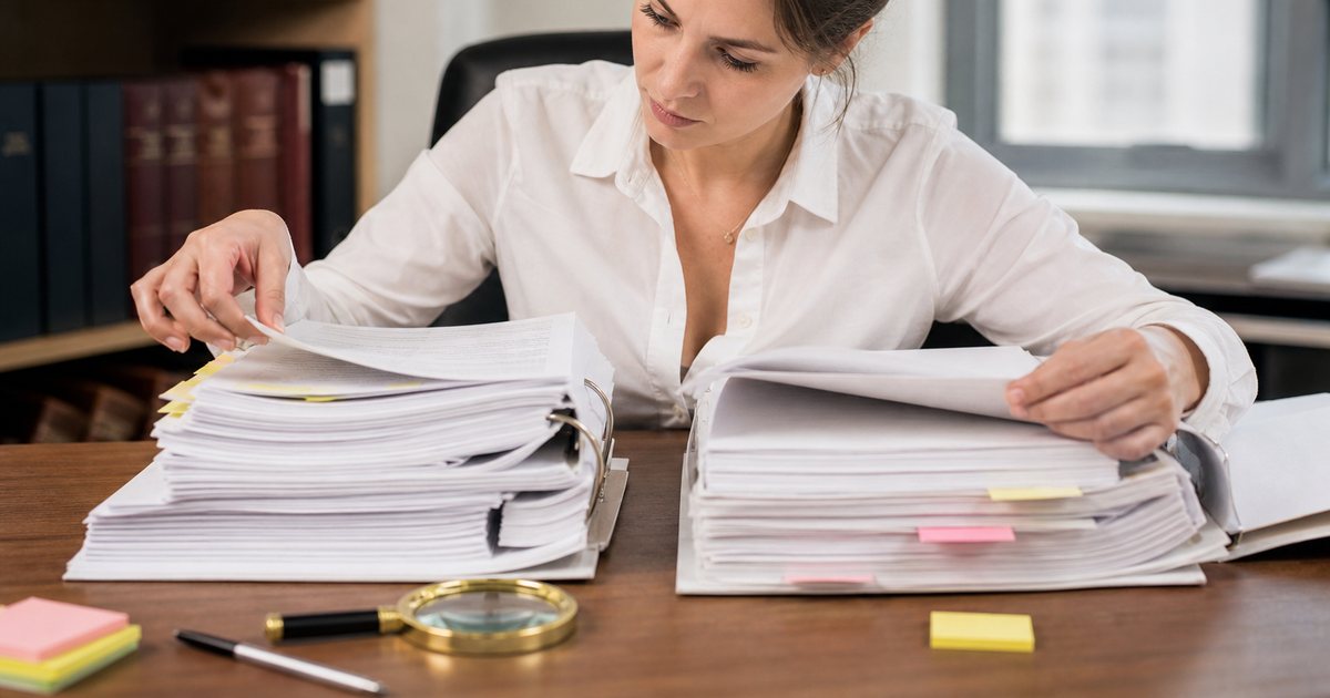 A paralegal comparing two thick printed binders side by side on a desk, flipping through tabbed dividers, a magnifying glass and sticky notes nearby